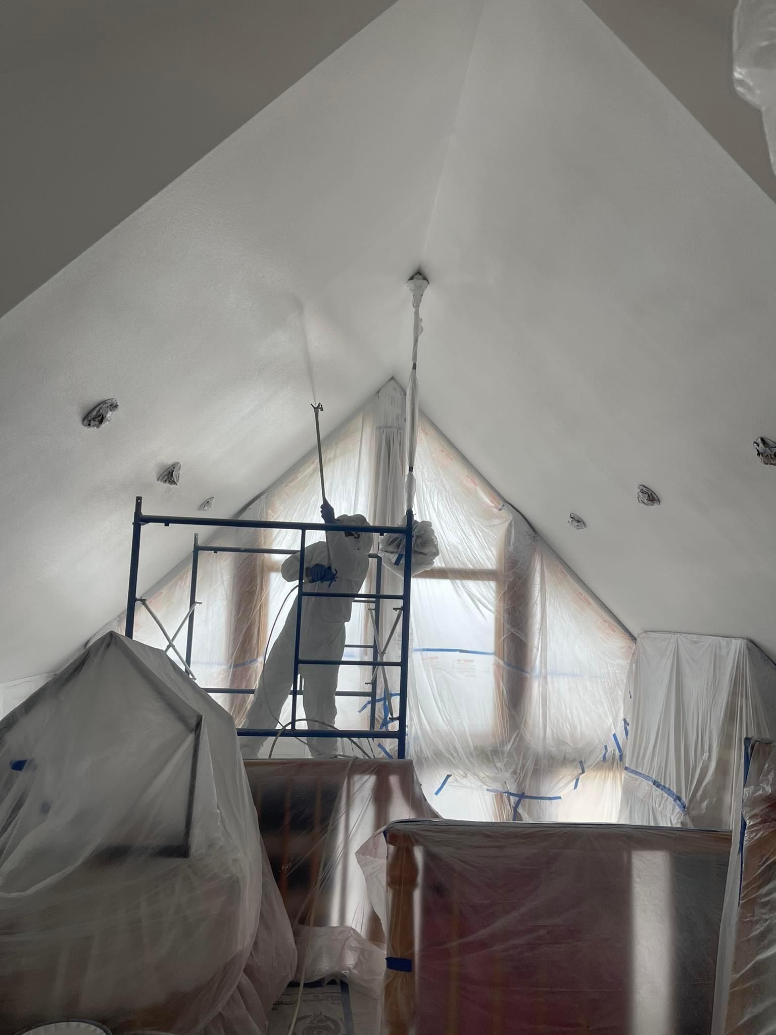 A man is standing on a scaffolding in an attic painting the ceiling.