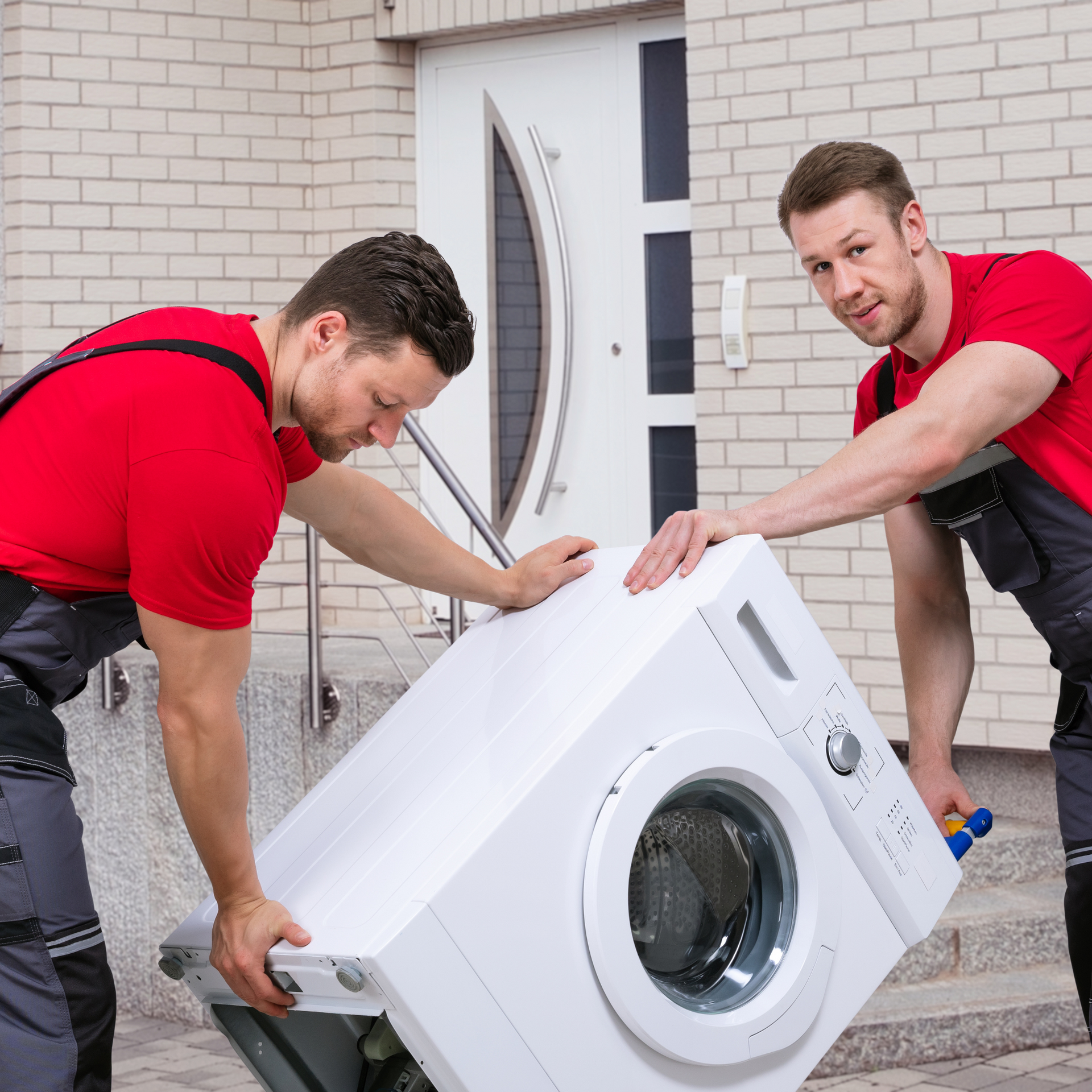 Two men in red shirts and gray overalls moving a white washing machine near a front door.
