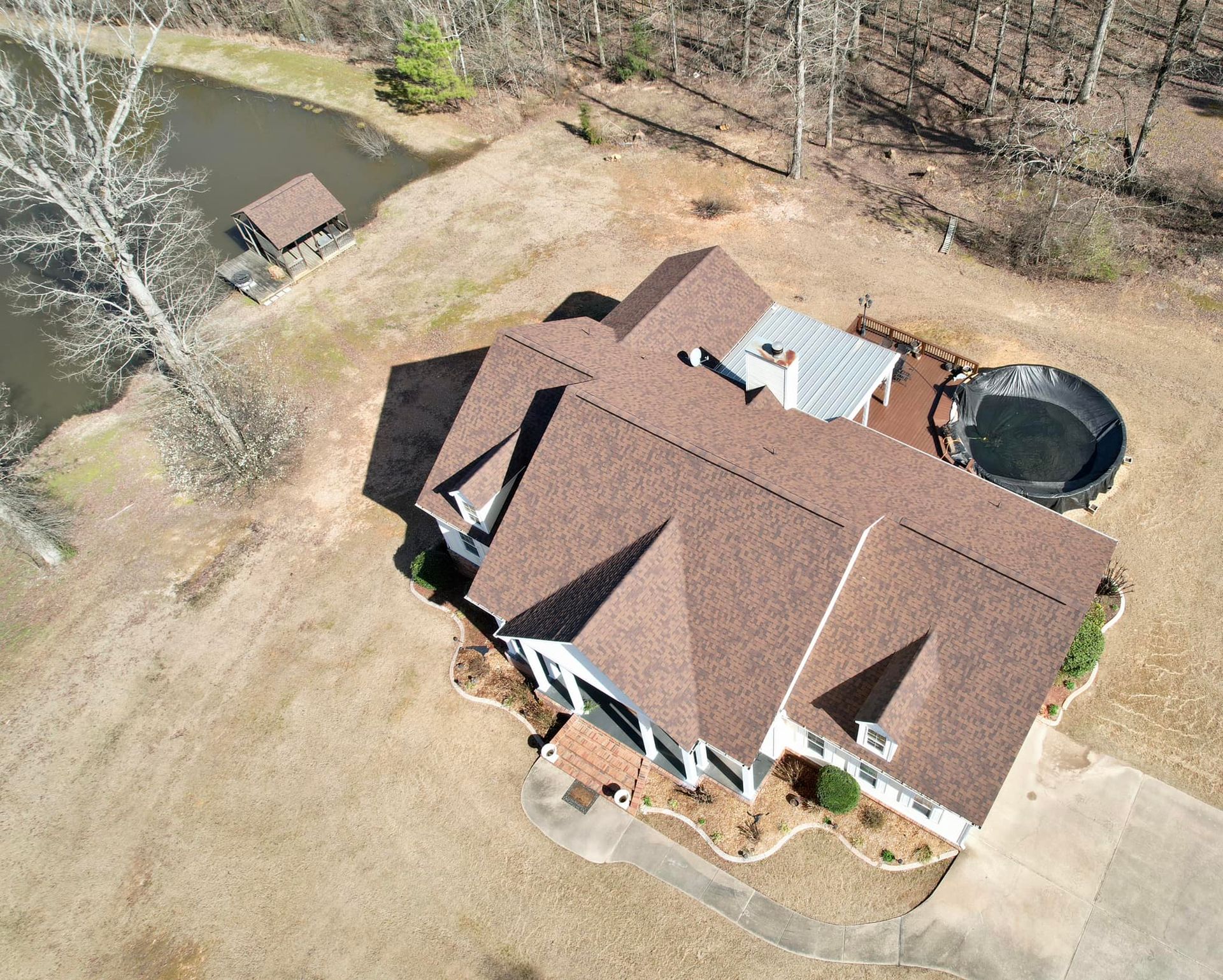 Aerial view of a house with a brown roof near a pond and a black above-ground pool.