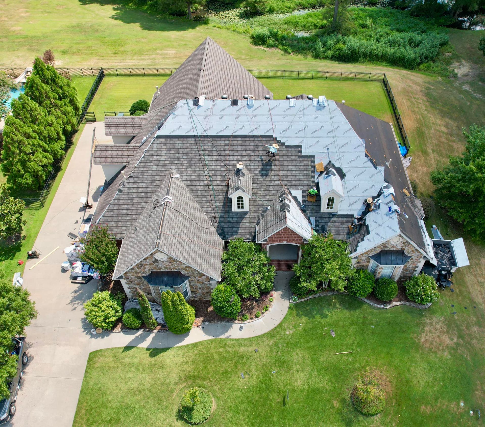 Aerial view of a house with a partially replaced roof; green lawn, trees, and swimming pool visible.