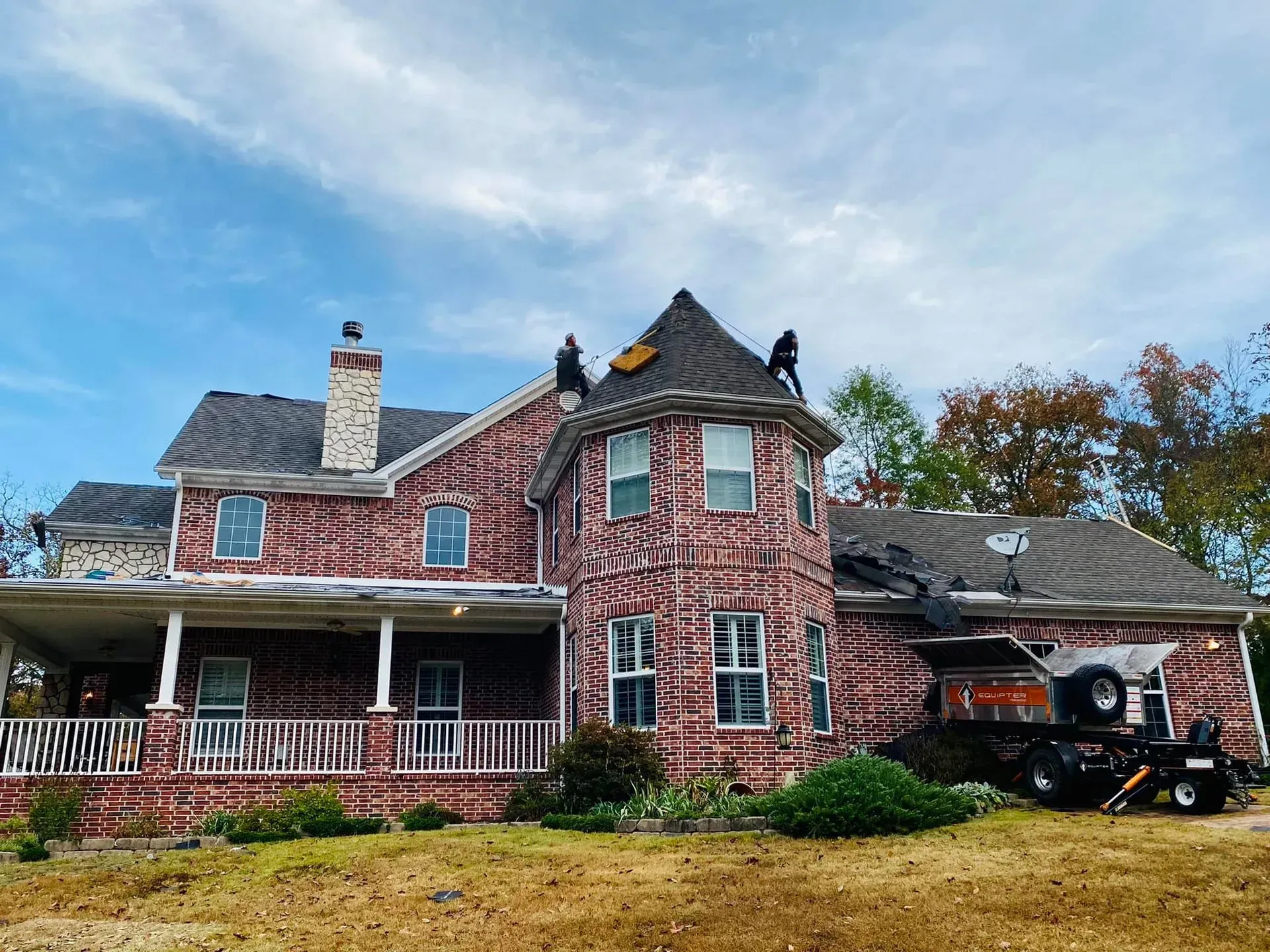 Brick house with workers on the roof, roof tear off in progress. A trailer with a debris chute is present.