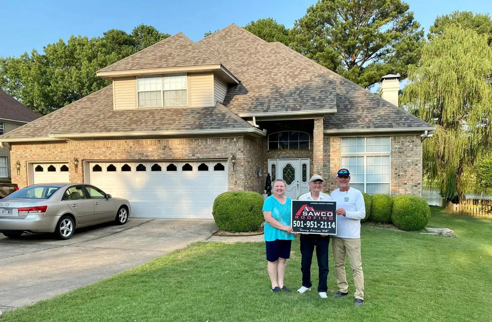People holding a sign in front of a brick house with a new roof and car in the driveway.