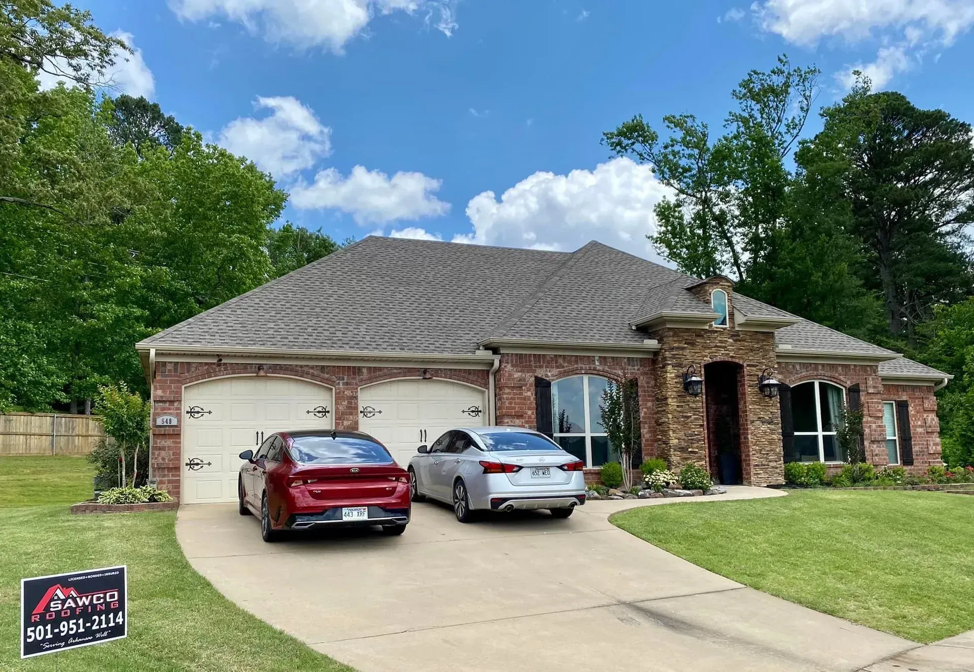 Brick house with two-car garage, two cars parked in the driveway, and a sign on the lawn.