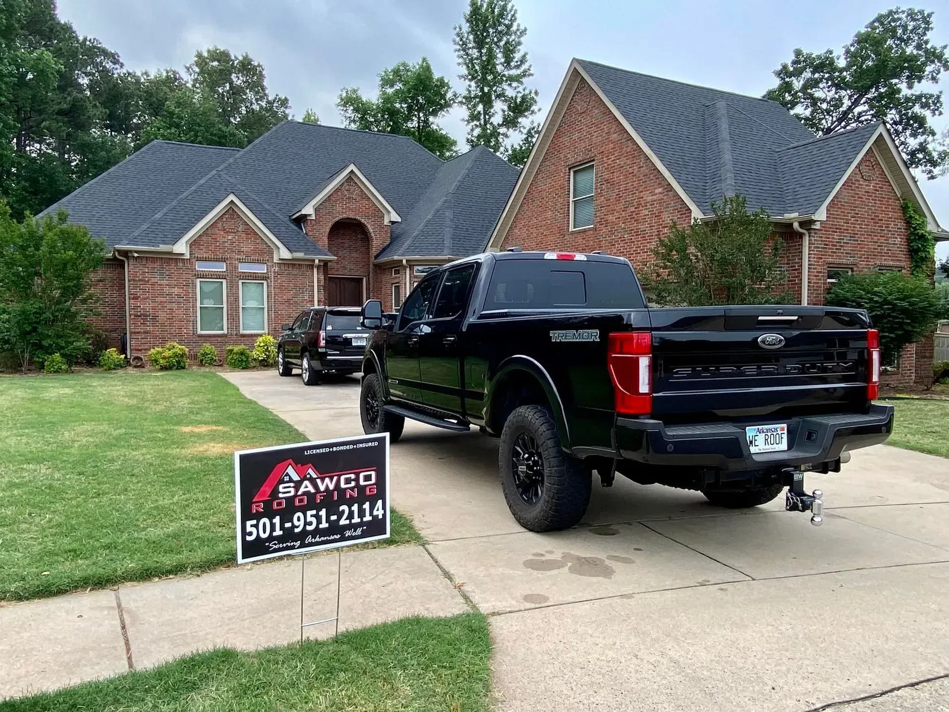 Black truck parked in front of a brick house with a sign for SAWED roofing. Green lawn, overcast sky.