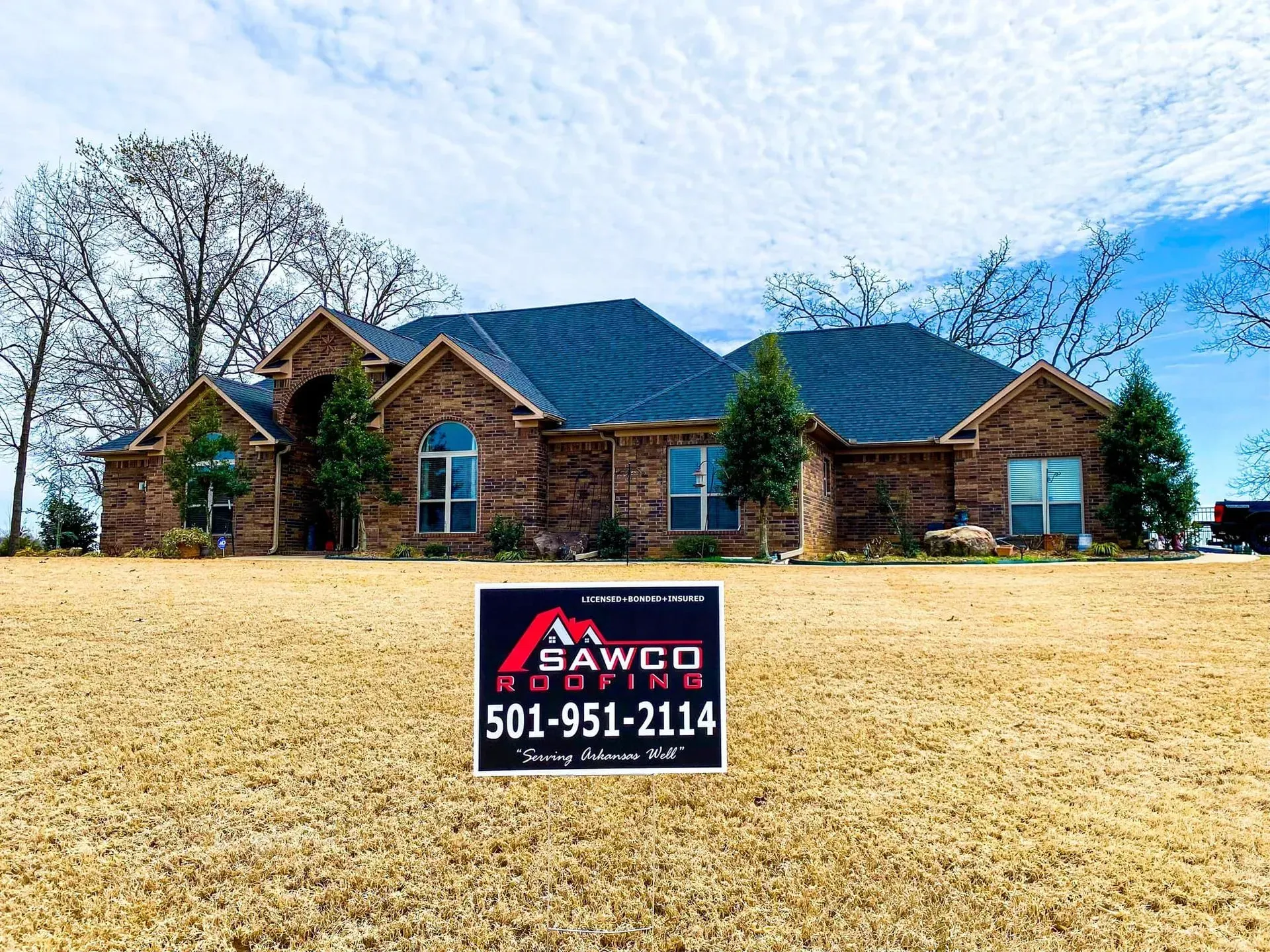 A sign for SAWCO Roofing in front of a brick house with a blue roof on a sunny day.
