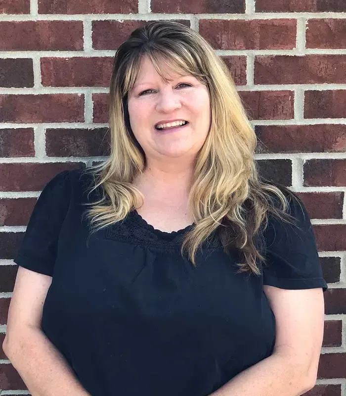 A woman in a black shirt is smiling in front of a brick wall.