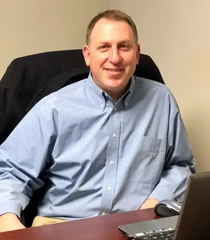 A man in a blue shirt is sitting at a desk with a laptop