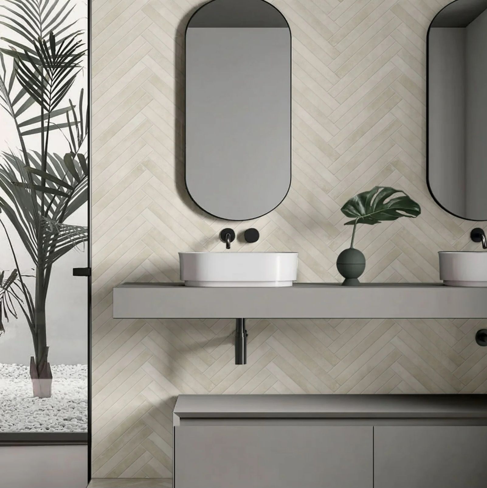 Modern bathroom with gray cabinets, a floating shelf with sinks, oval mirrors, and herringbone patterned light beige tile.