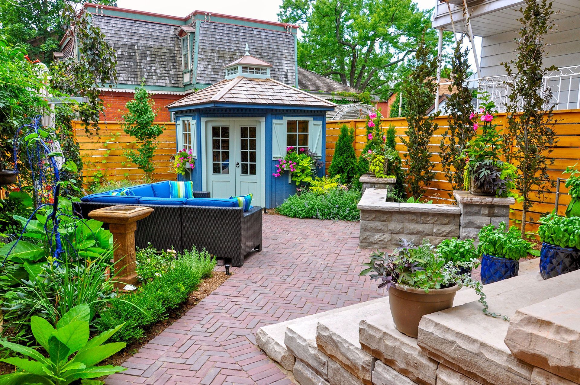 Colorful backyard patio with brick path, blue shed, potted plants, and hot tub in a landscaped garden