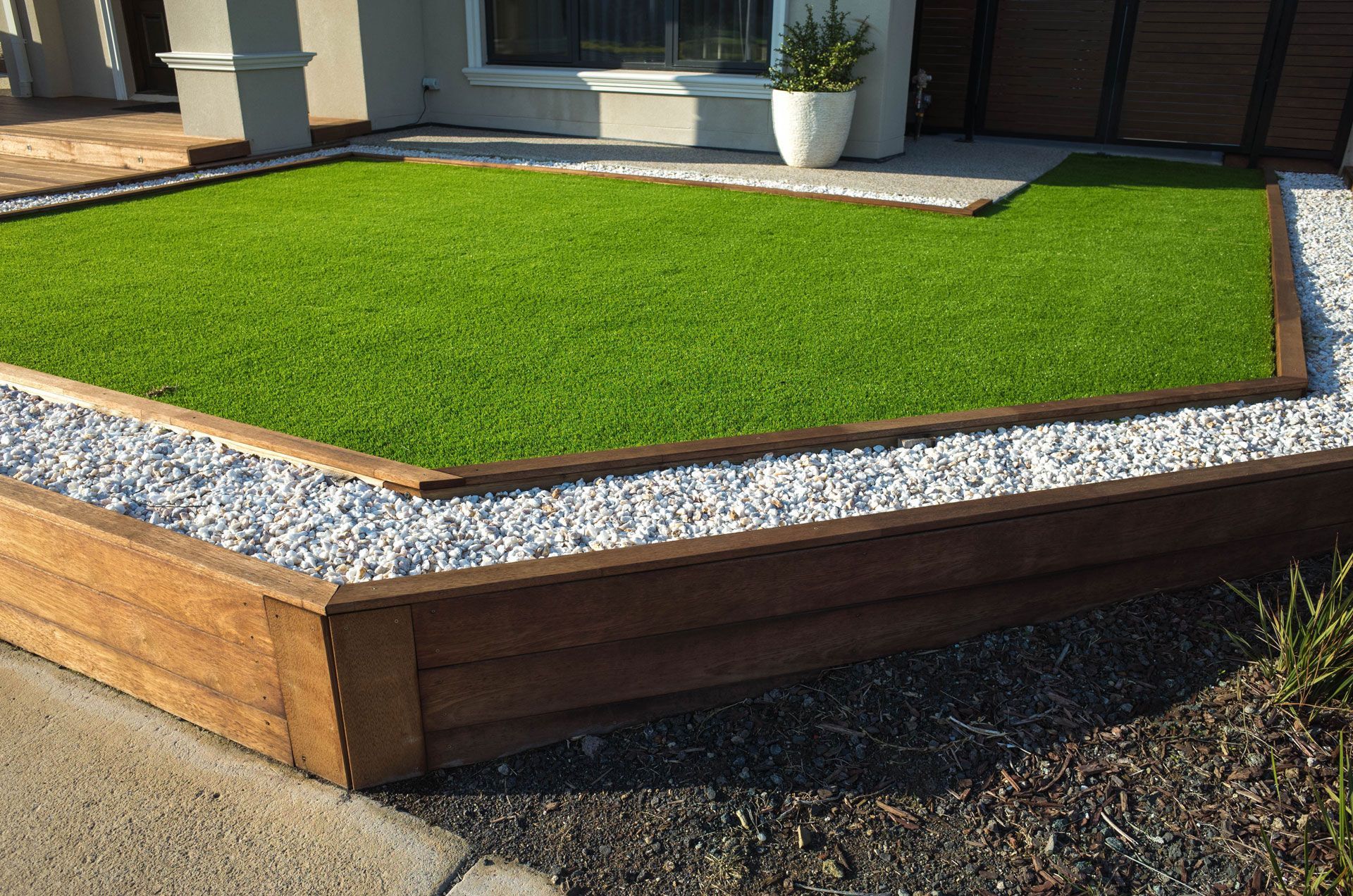 Raised artificial turf lawn bordered by wooden edging and white gravel beside a house.