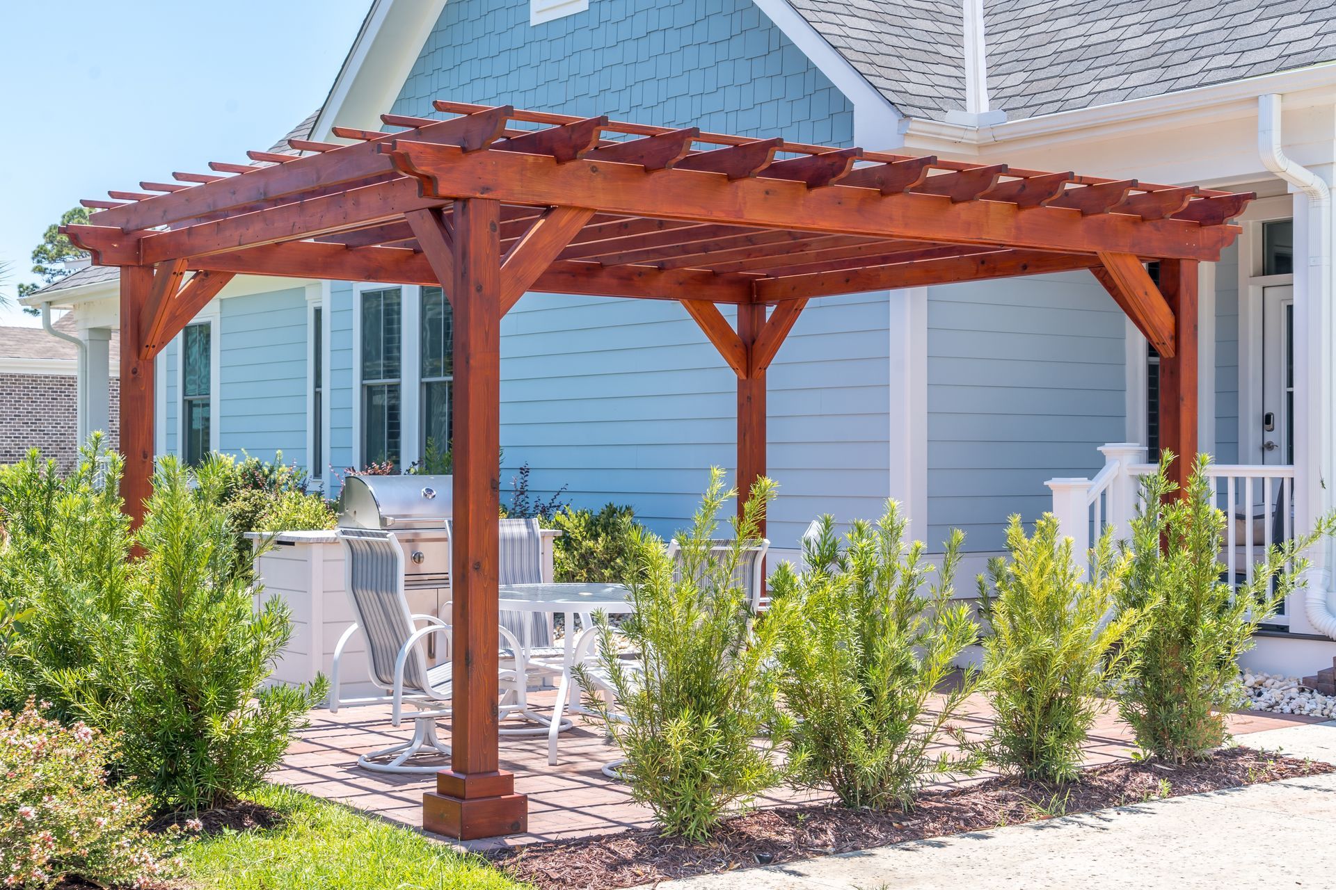 Wooden pergola over white patio furniture beside a blue house and garden shrubs