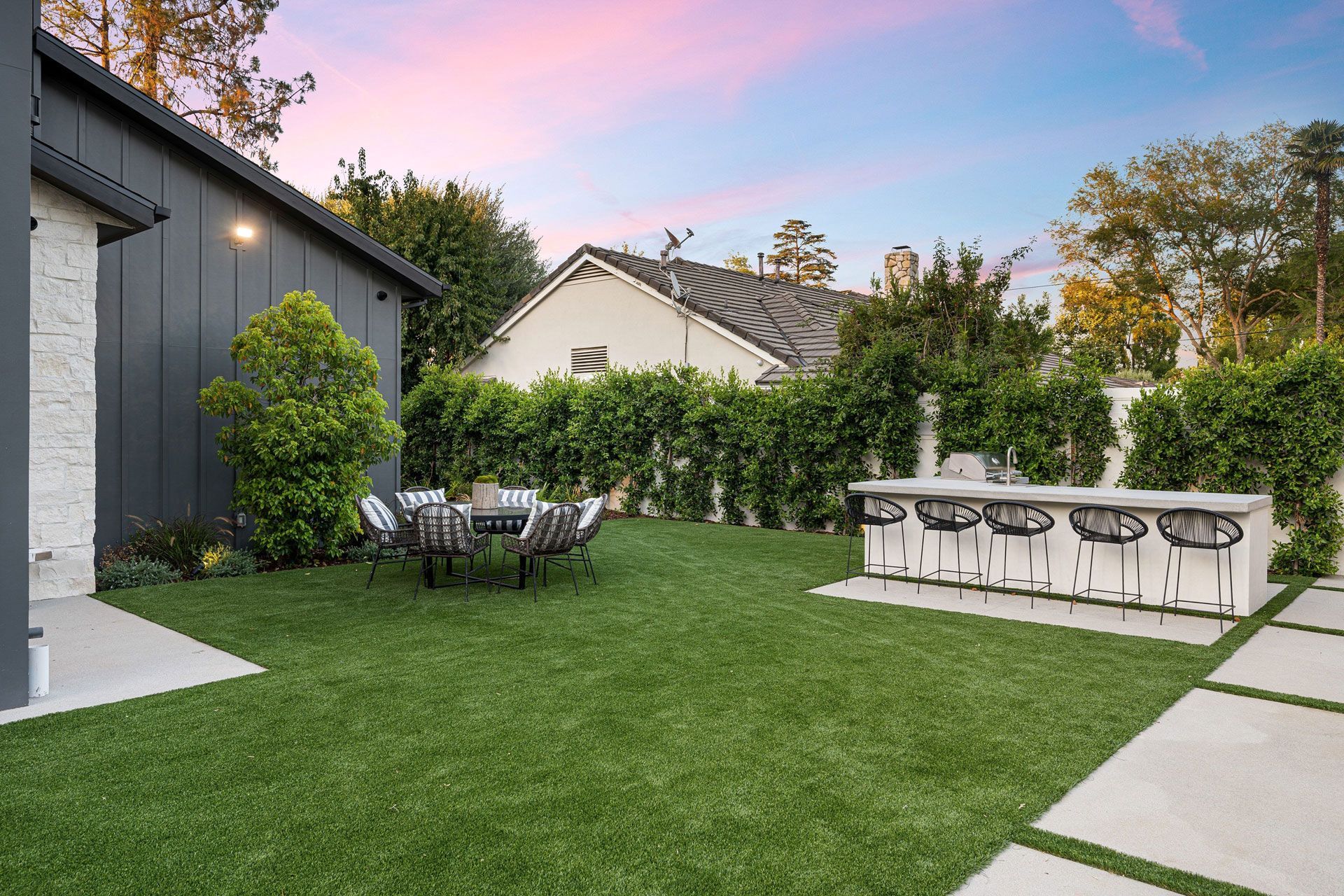 Modern backyard patio with lawn, hedge, dining table, and outdoor counter at sunset