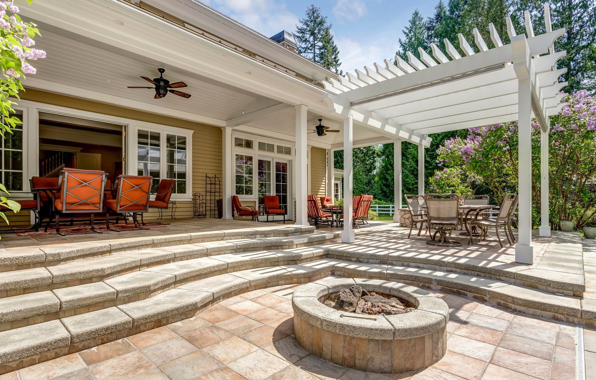 Patio with outdoor seating, white pergola, stone steps, and circular fire pit in a sunny backyard