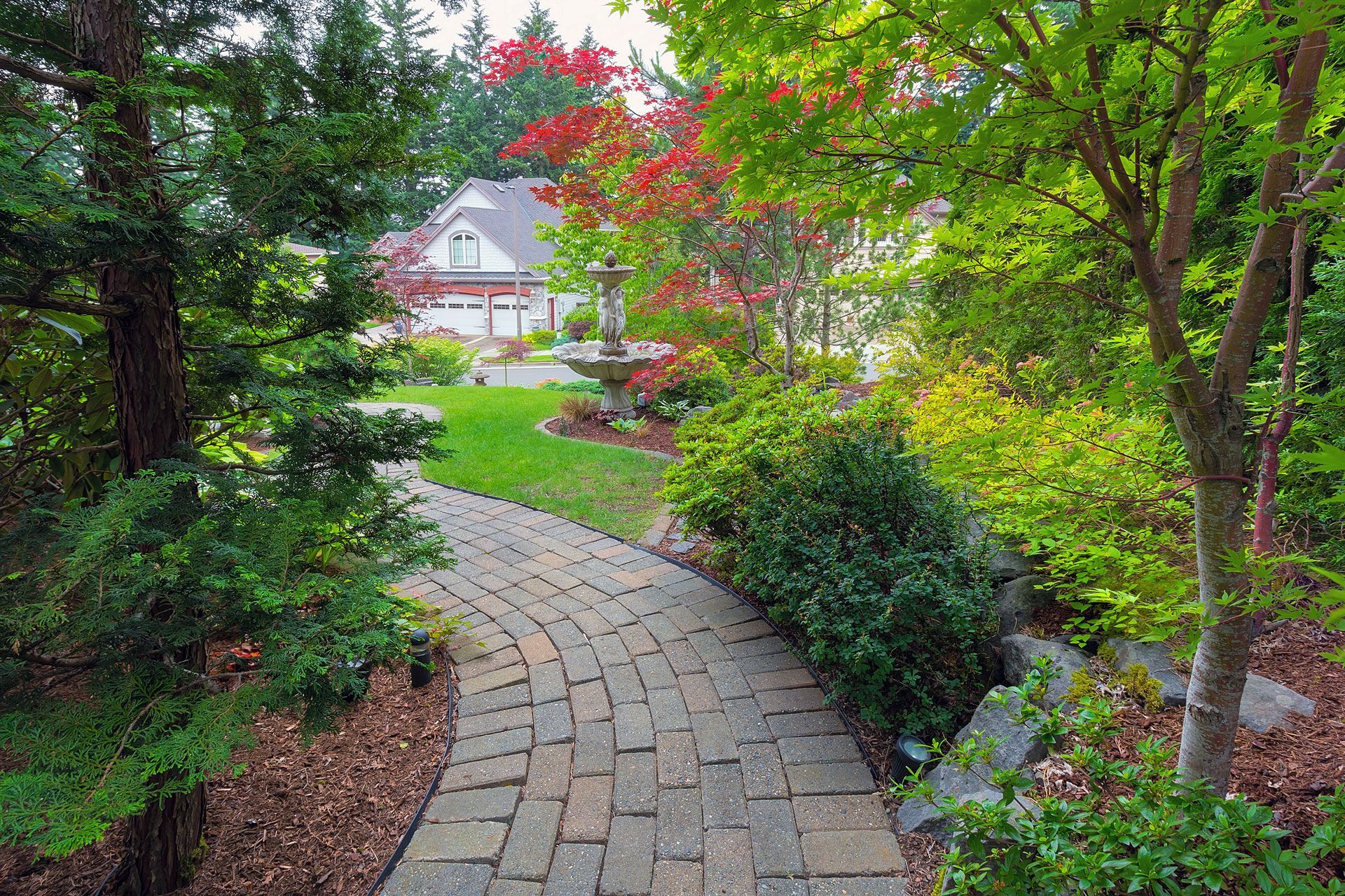 Curved stone path through lush garden toward a house, with red flowers and trees