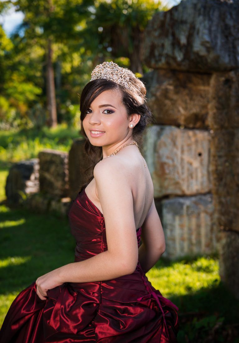 Young person in a formal maroon dress and tiara, smiling, outdoors near stone structure.
