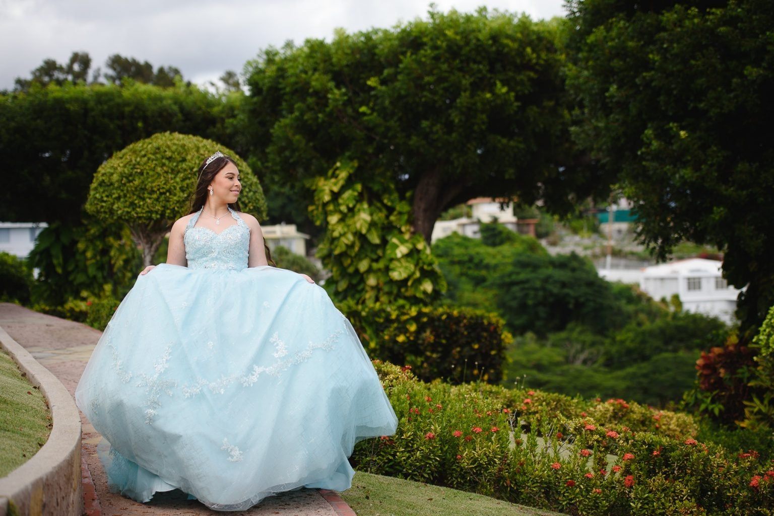 Woman in a light blue gown poses outdoors; trees and a stone walkway are in the background.
