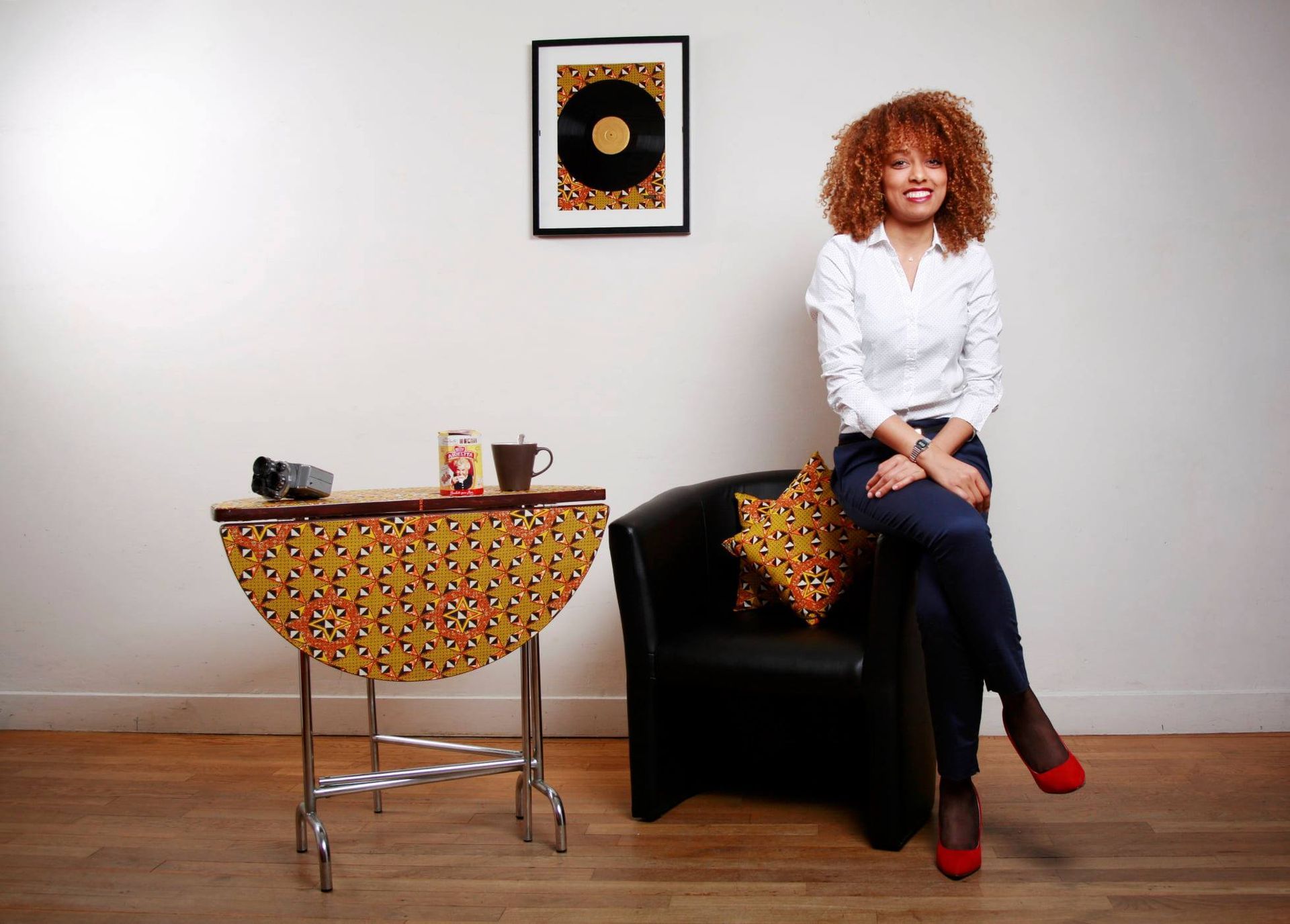 Woman in white shirt and jeans sits beside a patterned table and black chair, near a wall with art.
