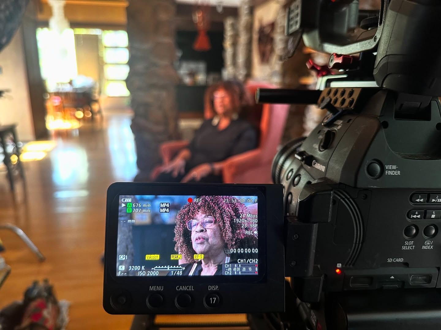 Camera filming a person sitting in a chair. The camera's screen shows the subject, who is in an indoor setting.