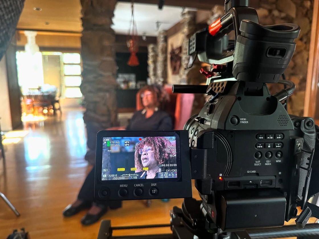 Camera filming a person sitting indoors. The camera screen shows the person speaking, while a room is blurred in the background.