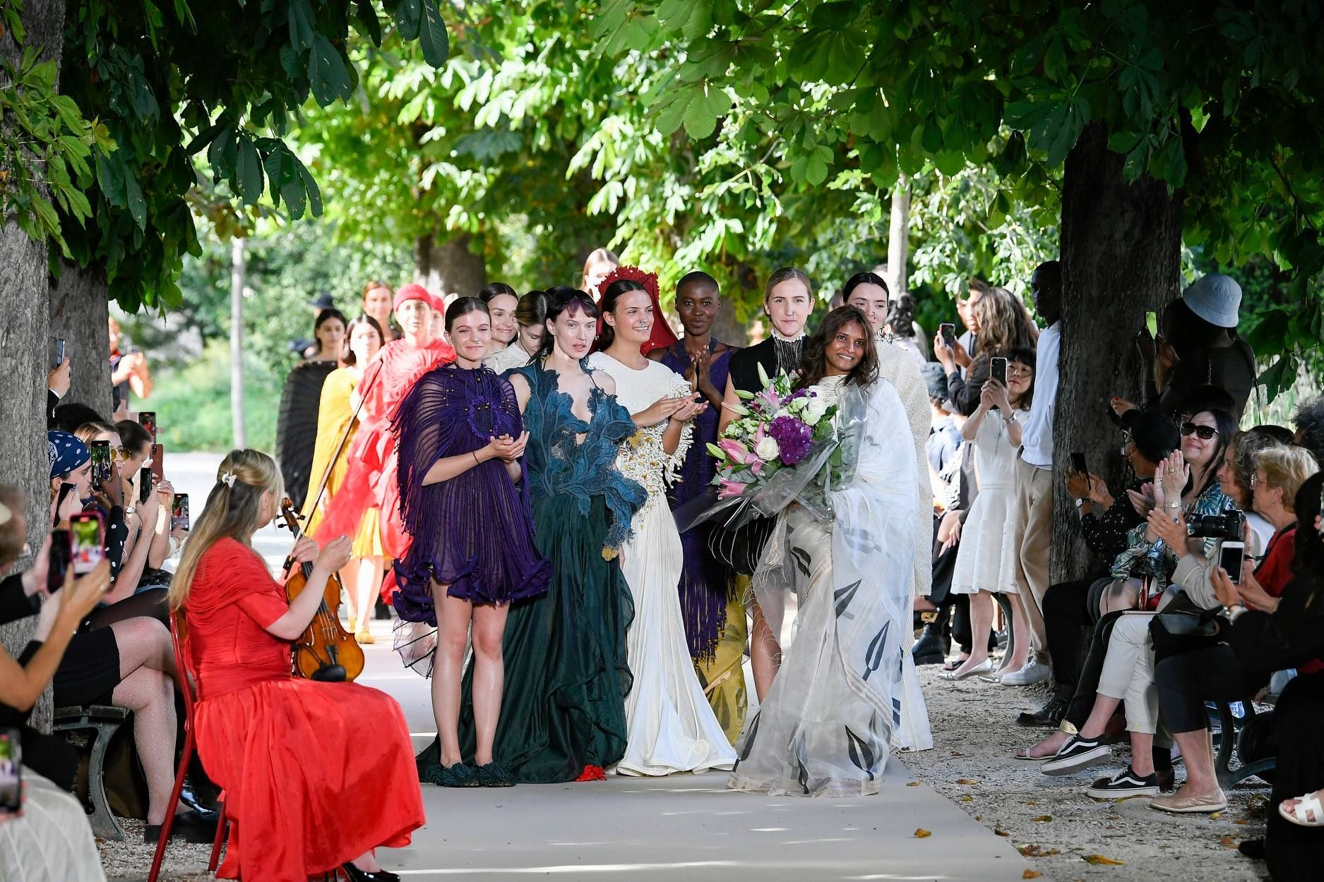 Models in colorful gowns on a runway outdoors, in front of an audience taking photos.