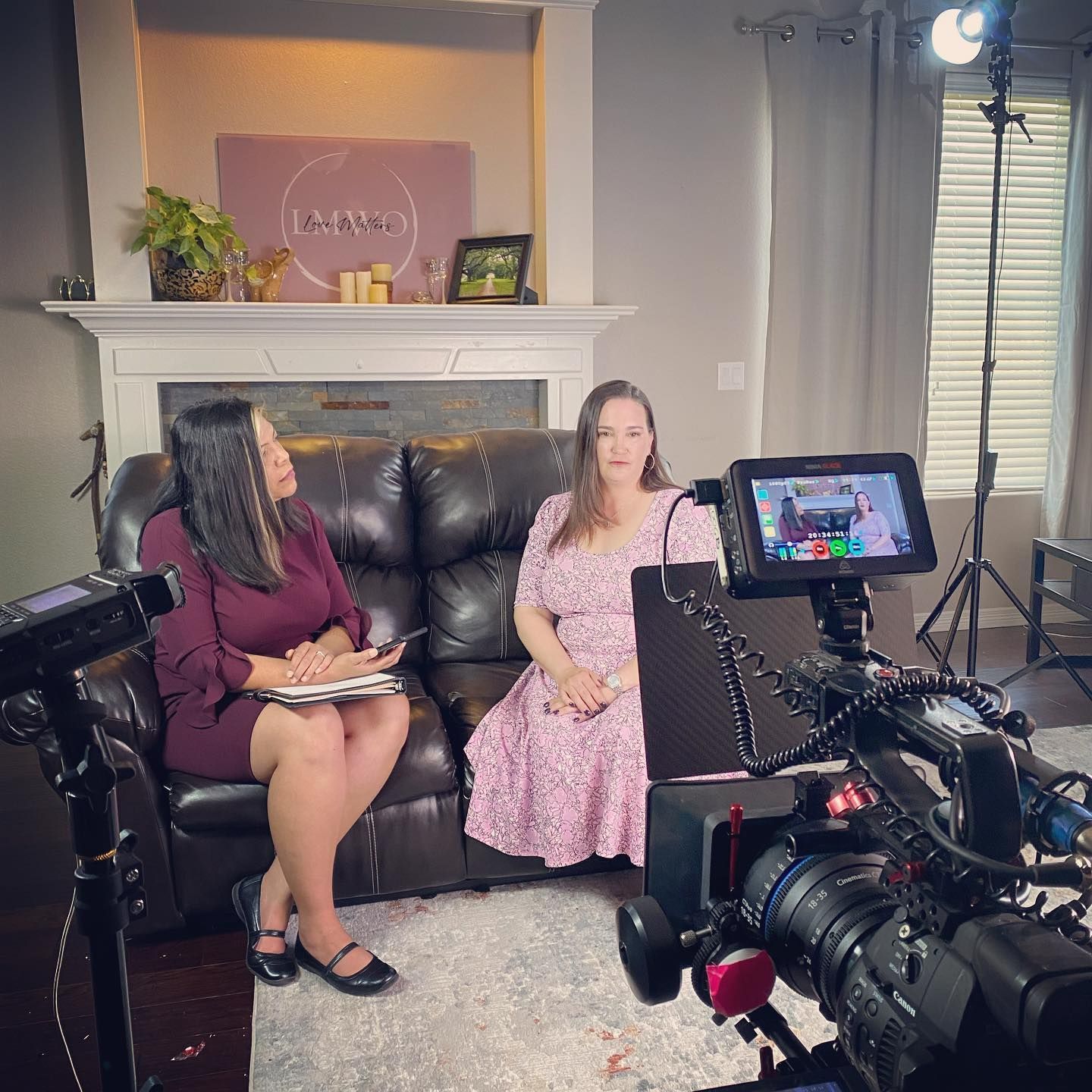 Two women on a couch being filmed. One holds a notepad. Cameras and lights are set up in a living room.