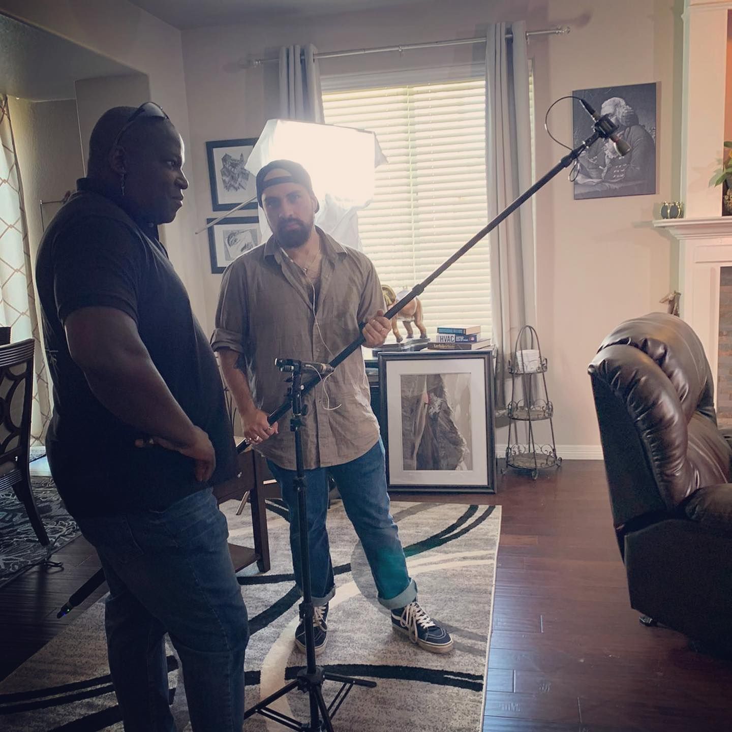 Two men filming inside a home; one holds a boom mic, the other stands. Sunlight streams from the window.