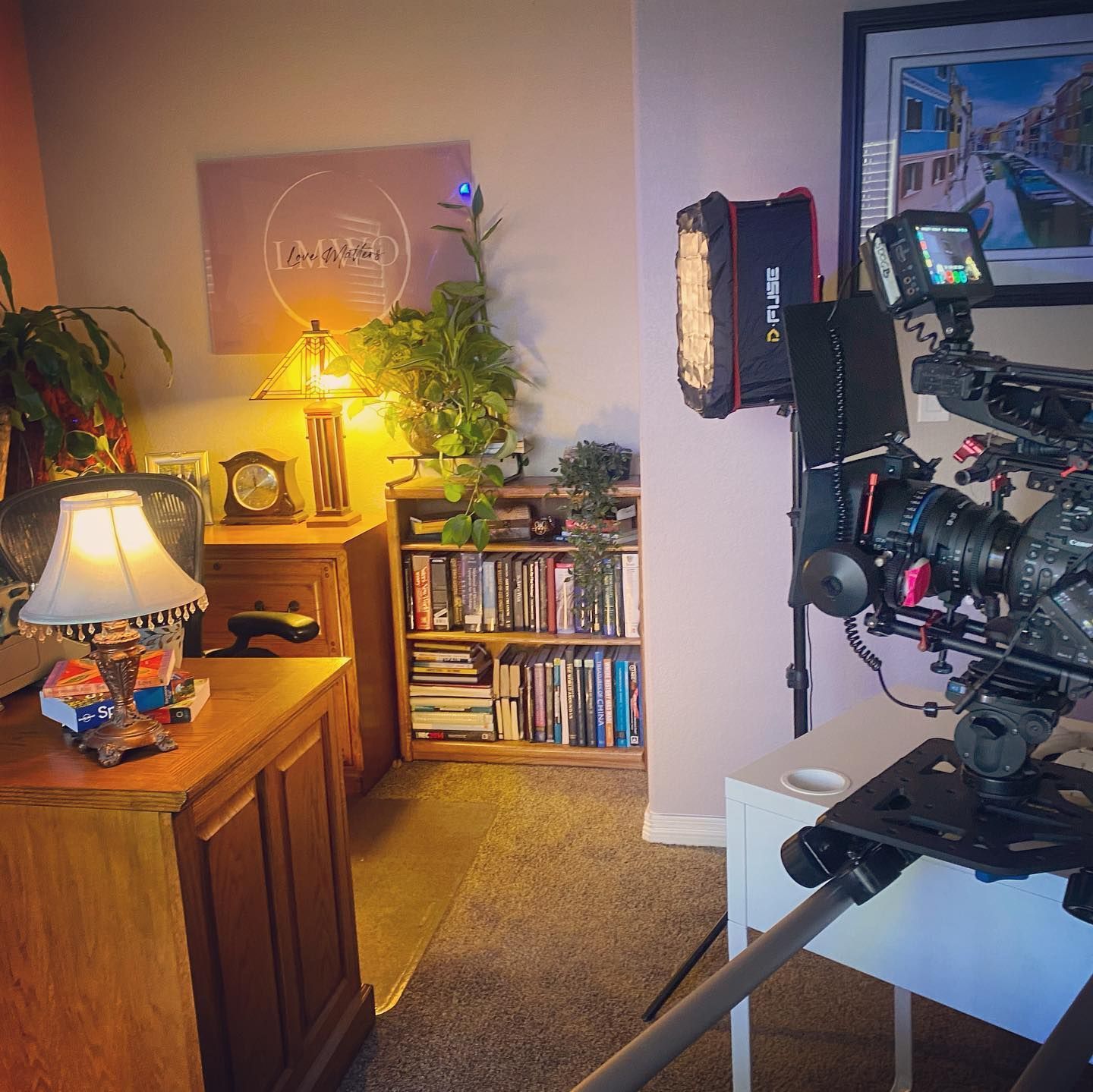 A film set with a camera on a tripod, lights, and a desk in a room.