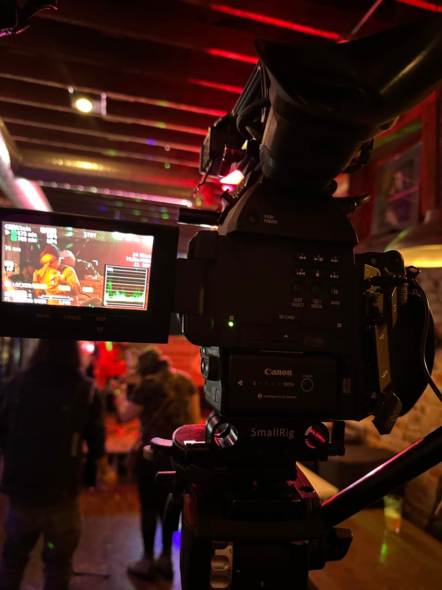 Camera on tripod filming in dimly lit room, with people and red lights in the background.