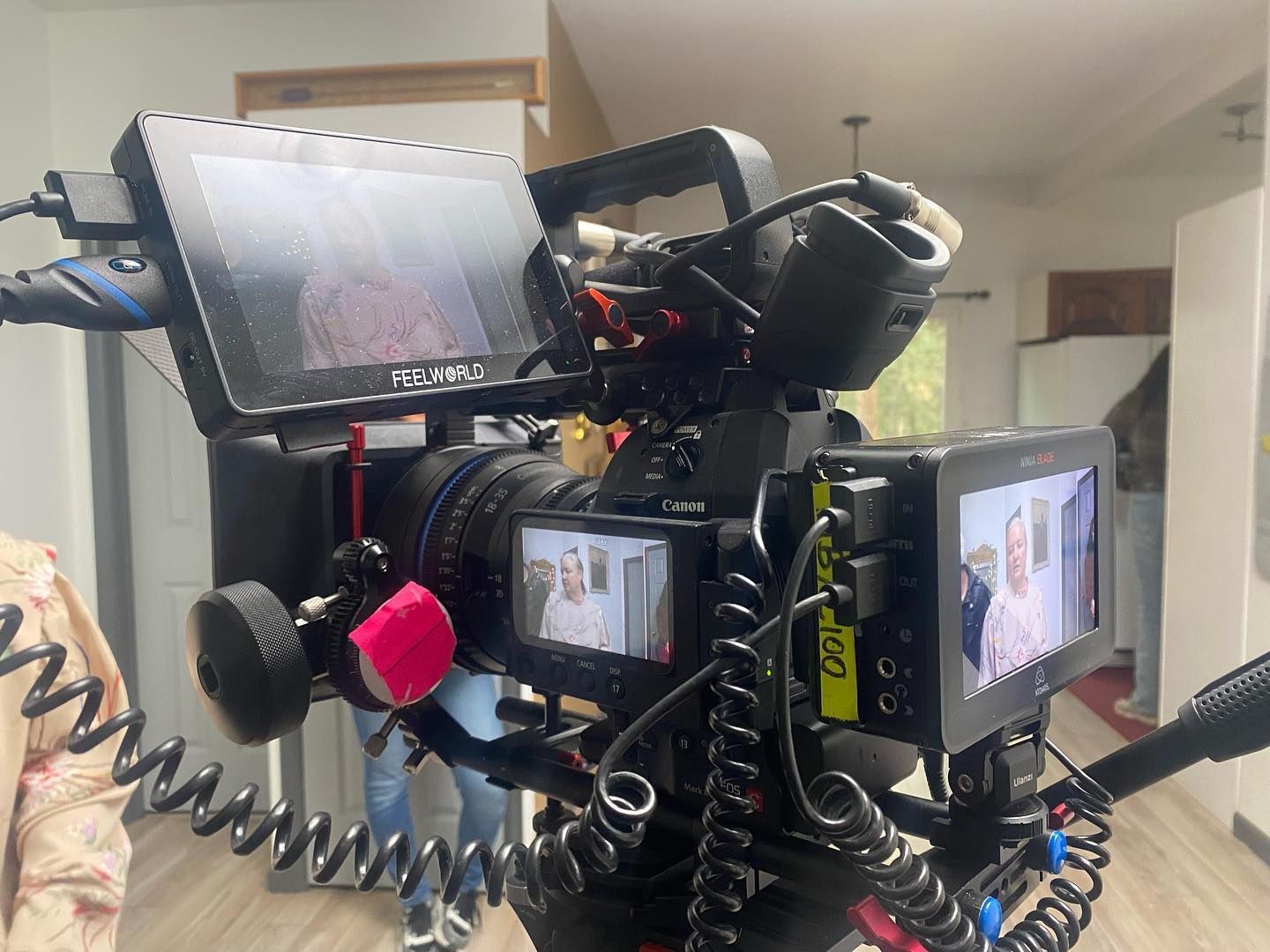Video camera on a tripod with multiple attached monitors; filming indoors.