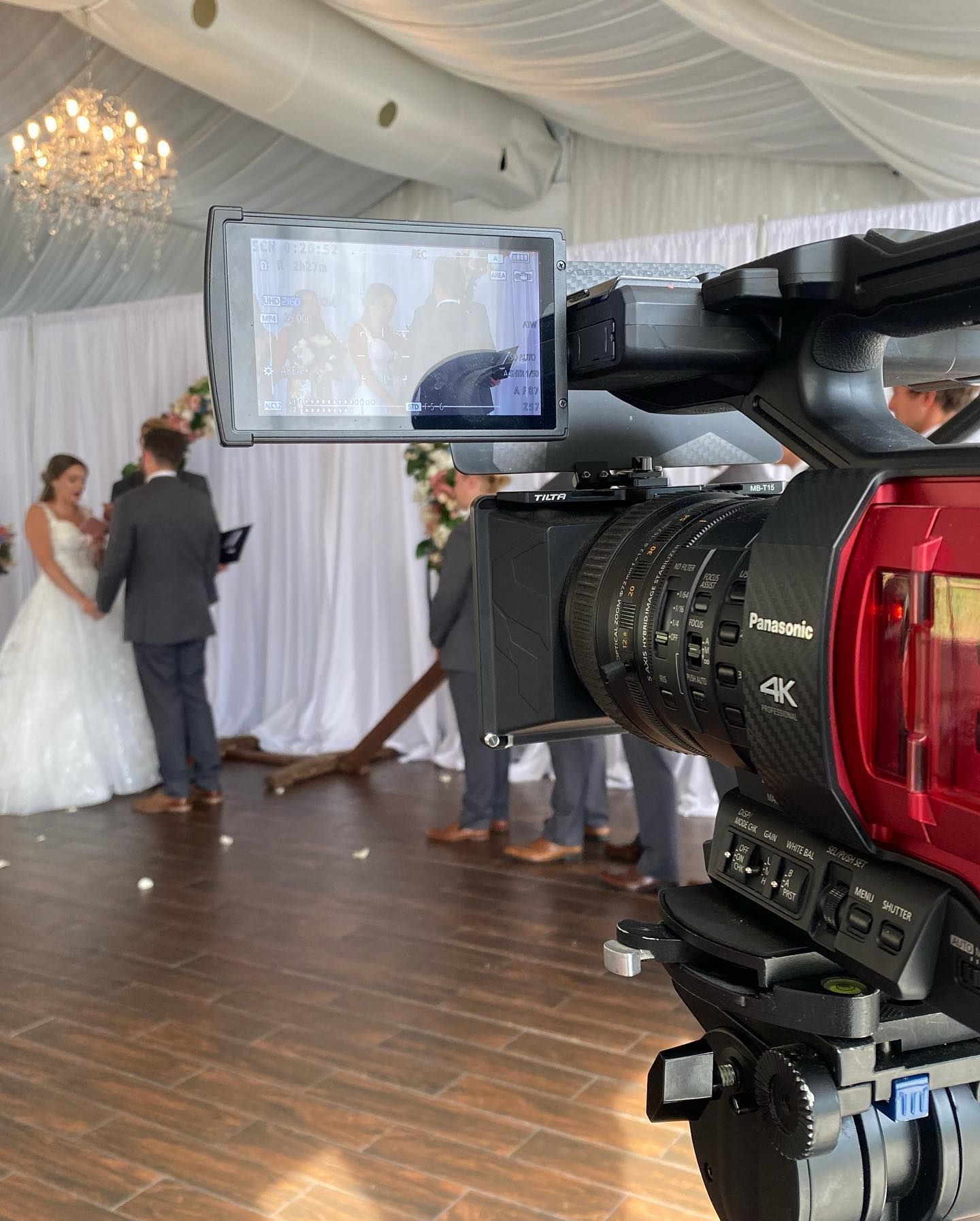 Wedding ceremony filmed with a professional video camera. Bride and groom stand before a wooden cross.