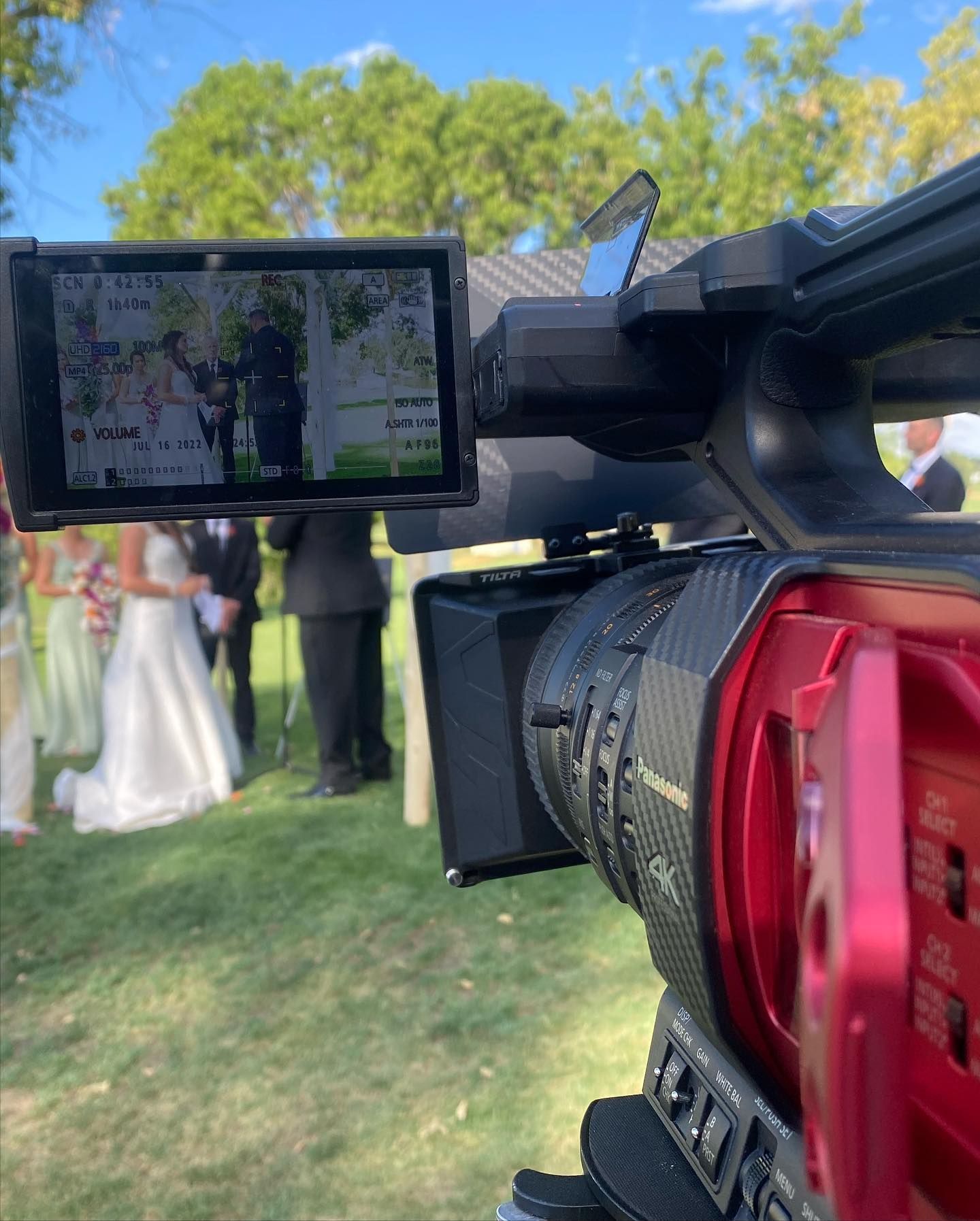 Video camera filming a wedding ceremony outdoors; bride, groom, and wedding party are visible on the monitor screen.