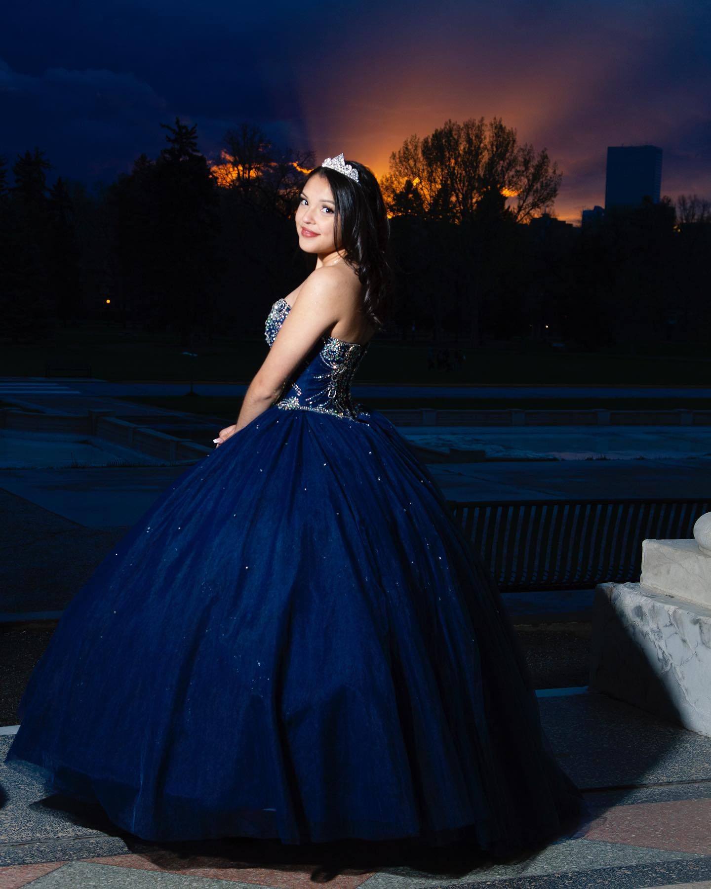 Woman in navy gown and tiara smiles outdoors at dusk.