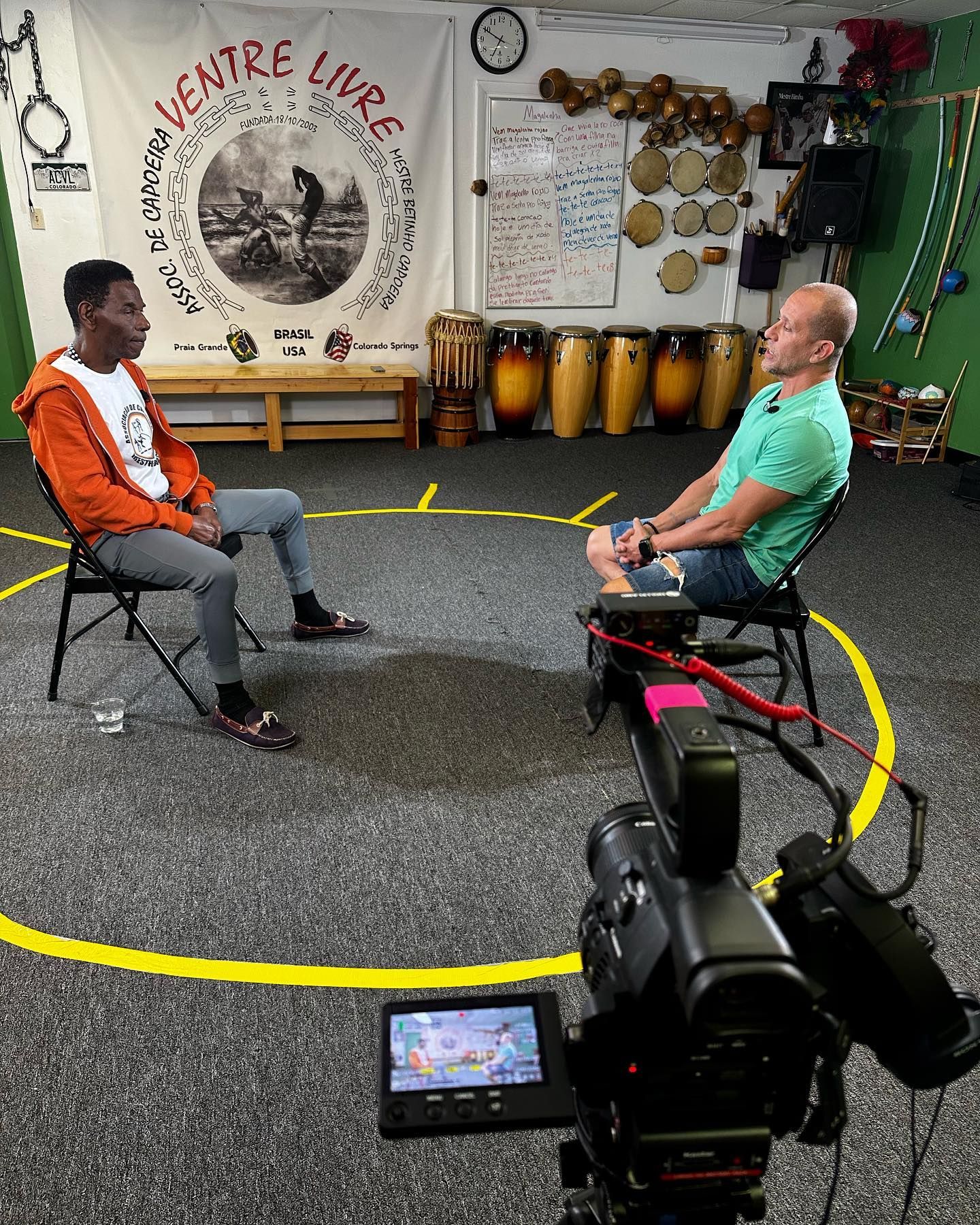 Two men in chairs are interviewed inside a studio. A video camera and a yellow circle are visible.