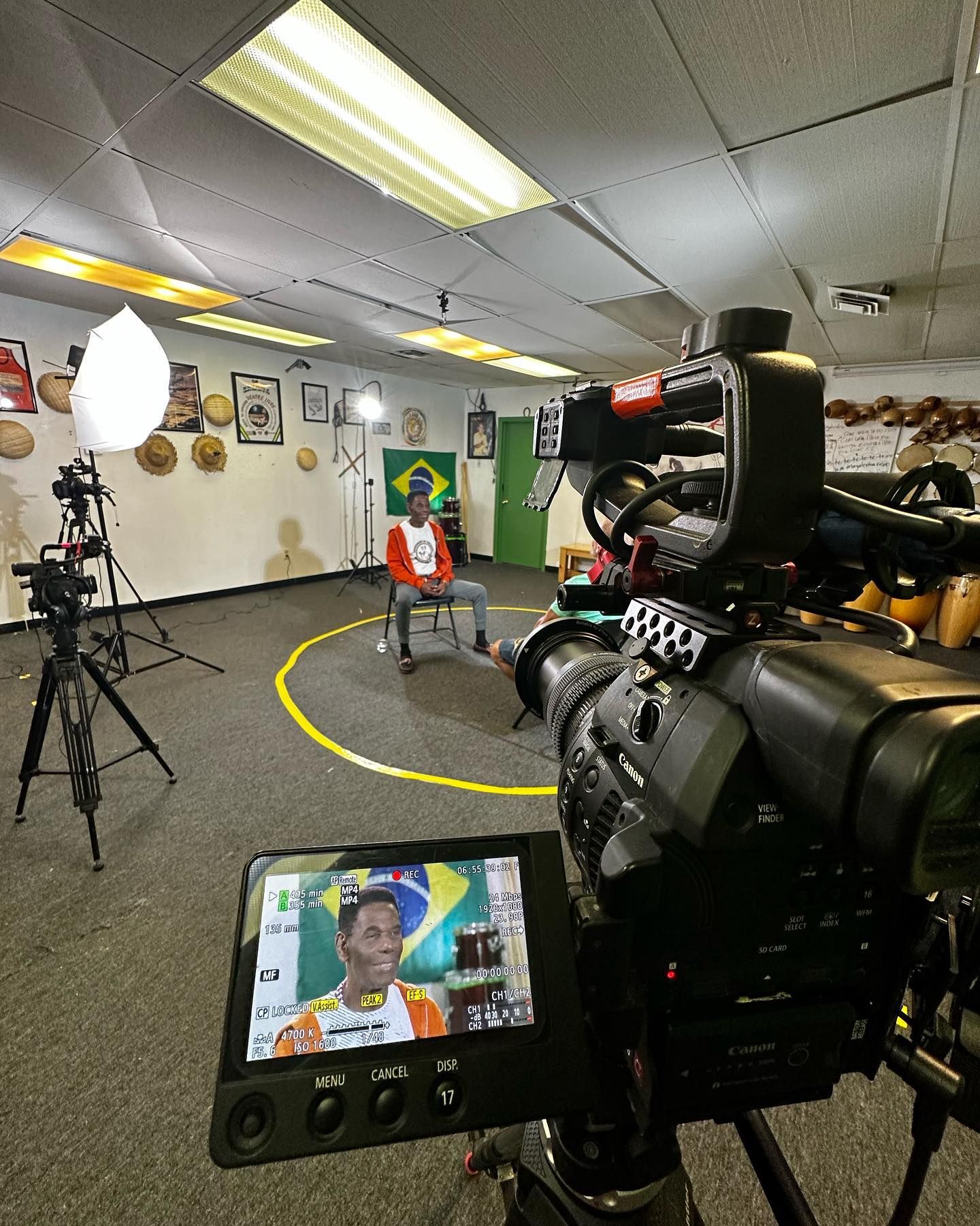 Video production set: a person is interviewed in a room. Cameras, lights, and crew are present.