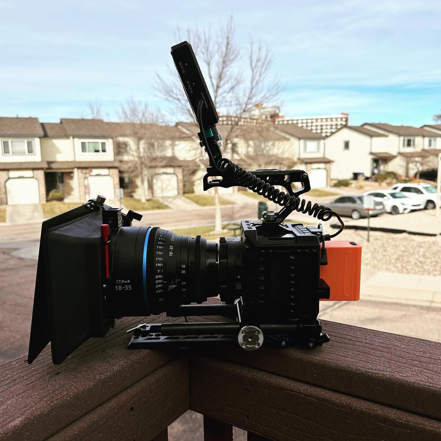 Black film camera with a microphone mounted, on a wooden railing, in front of townhouses and a bright sky.