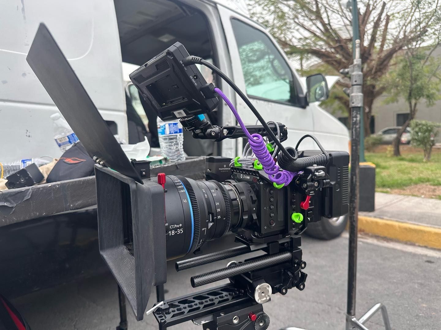 A professional film camera with a matte box and monitor is mounted on a tripod in front of a white van.