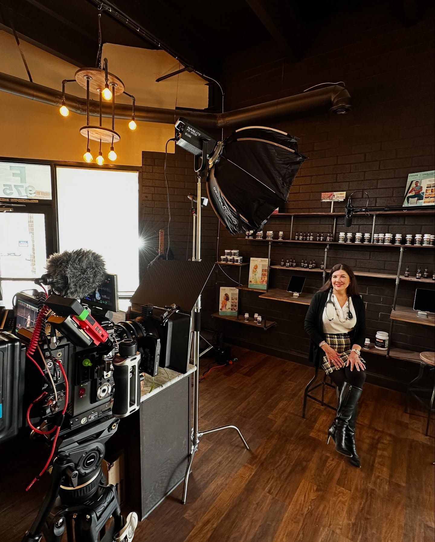 A woman sits on a stool being filmed in a store. Camera equipment and lights are set up.
