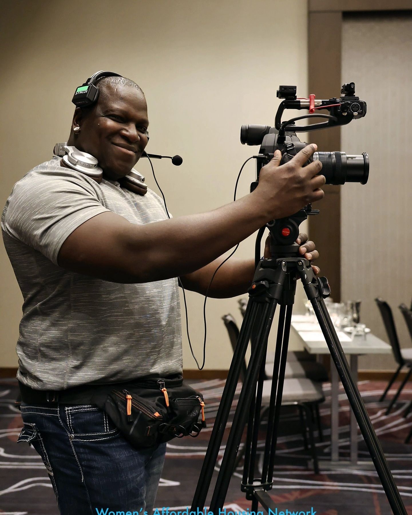 Videographer operating a camera on a tripod, wearing headphones and a fanny pack, smiling indoors.