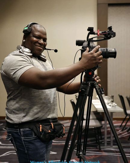 Person filming with a video camera on a tripod, smiling. Wearing a headset microphone, fanny pack, in a conference room.