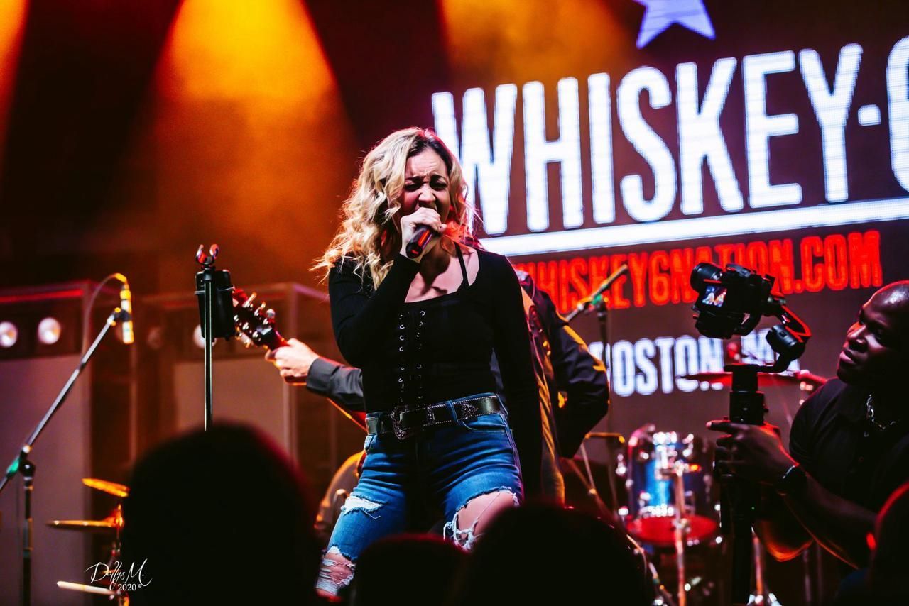 Woman singing on stage at Whiskey Grill, wearing black top and denim shorts, bathed in stage lights.