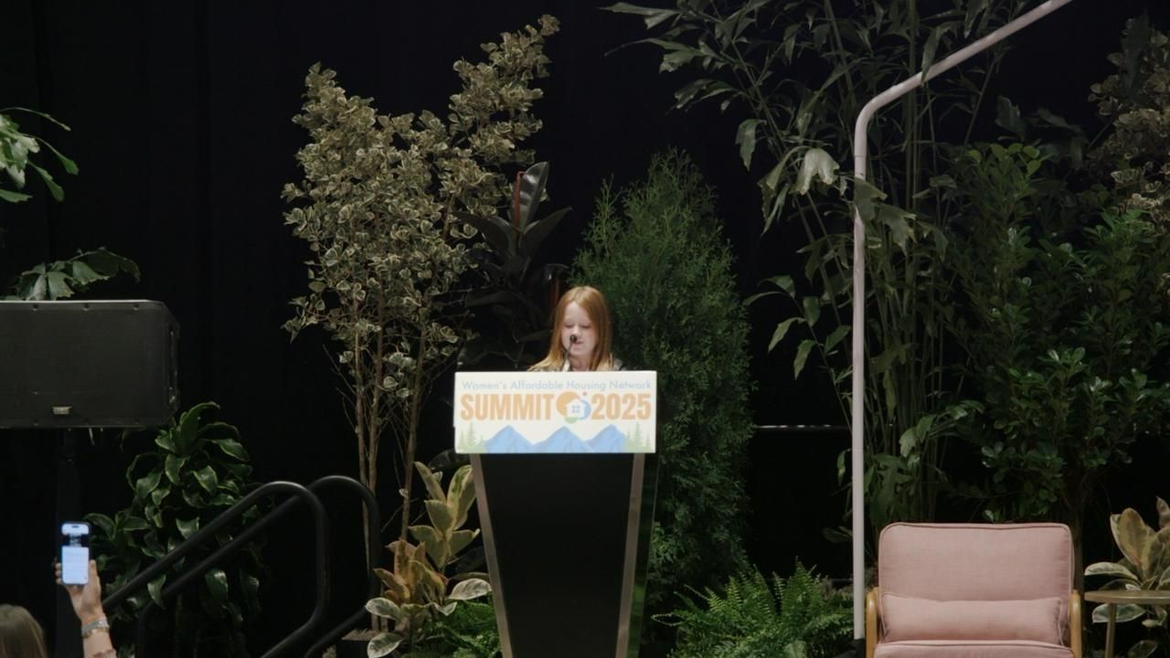 Young person speaking at a podium during an event. Plants and a pink chair are visible in the background.