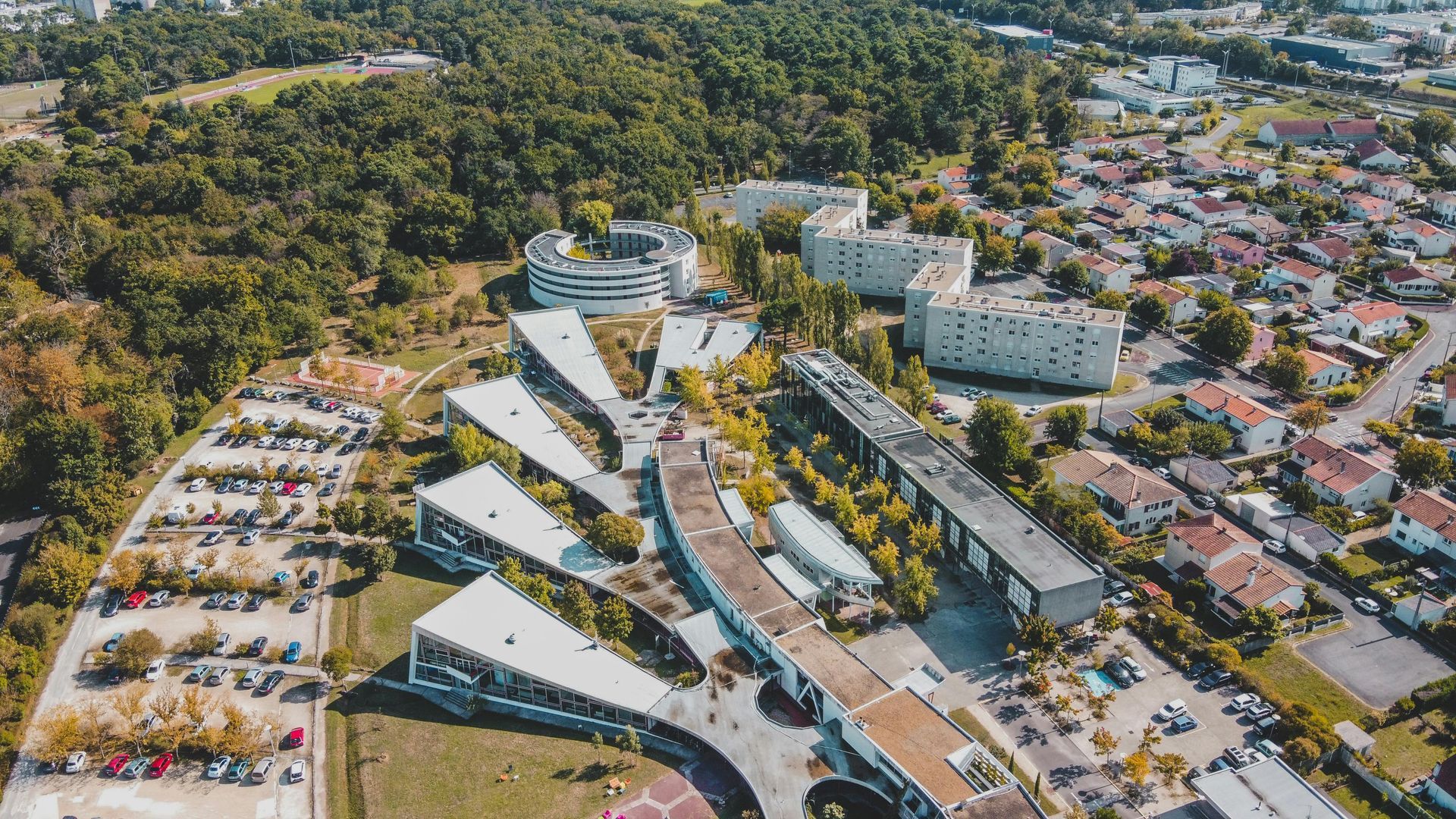 Aerial view of modern buildings with angular rooflines, a parking lot, and residential area, next to a wooded area.
