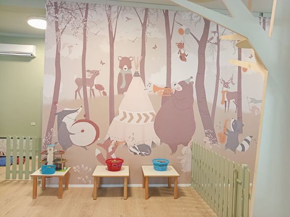 Sala giochi con murale raffigurante un bosco, tavolini e recinzione decorativa.