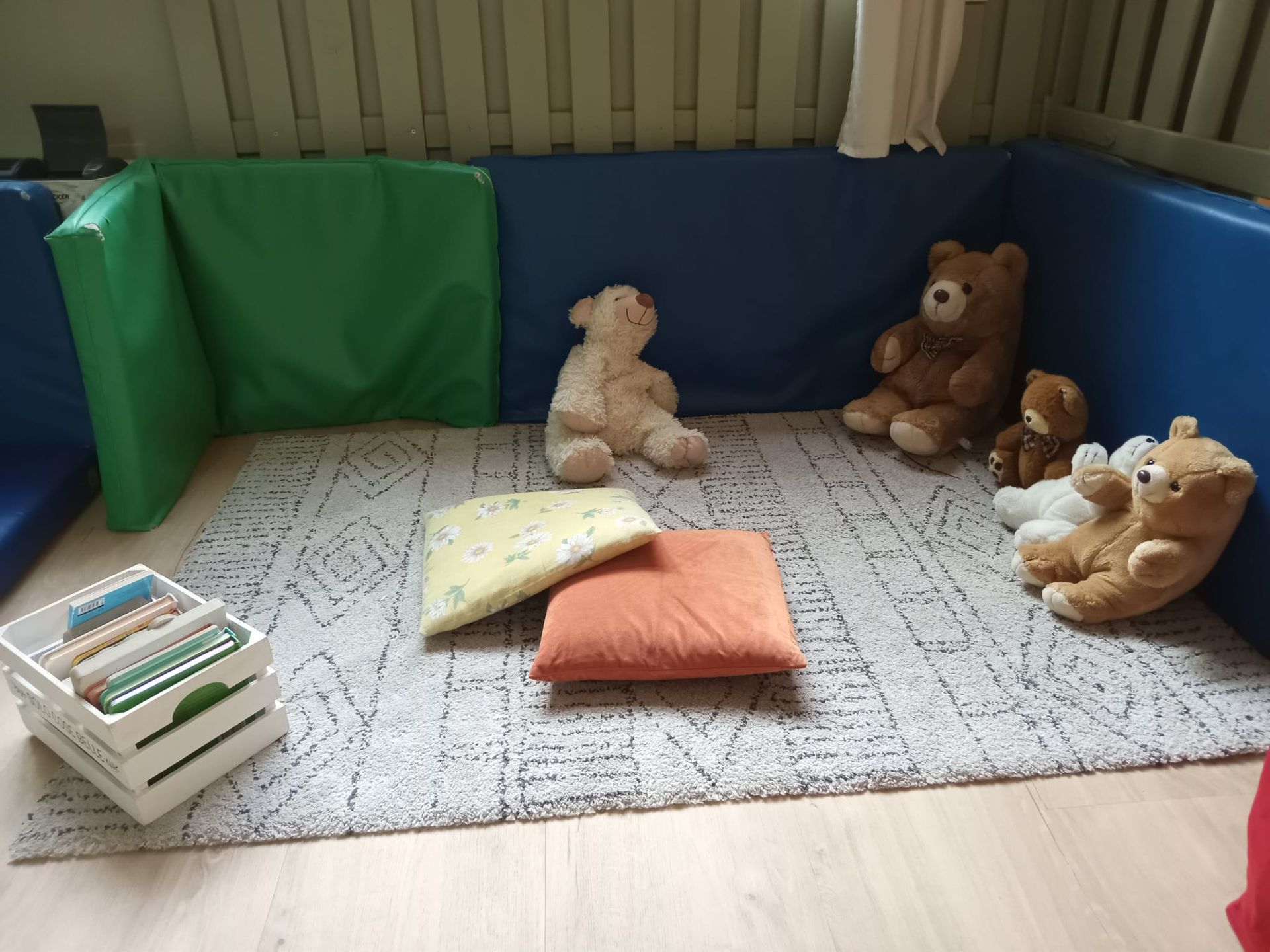 Accogliente area giochi con pareti imbottite blu e verdi, tappeto, orsacchiotti e cuscini. Cassa bianca con libri.