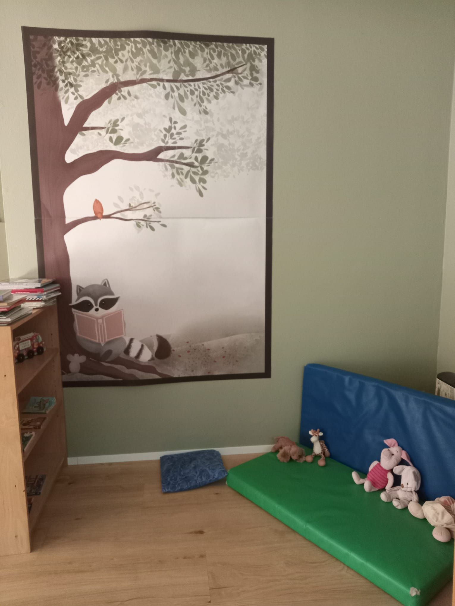 Un angolo lettura con disegni raffiguranti procioni, un tappetino verde, animali di peluche e una libreria.