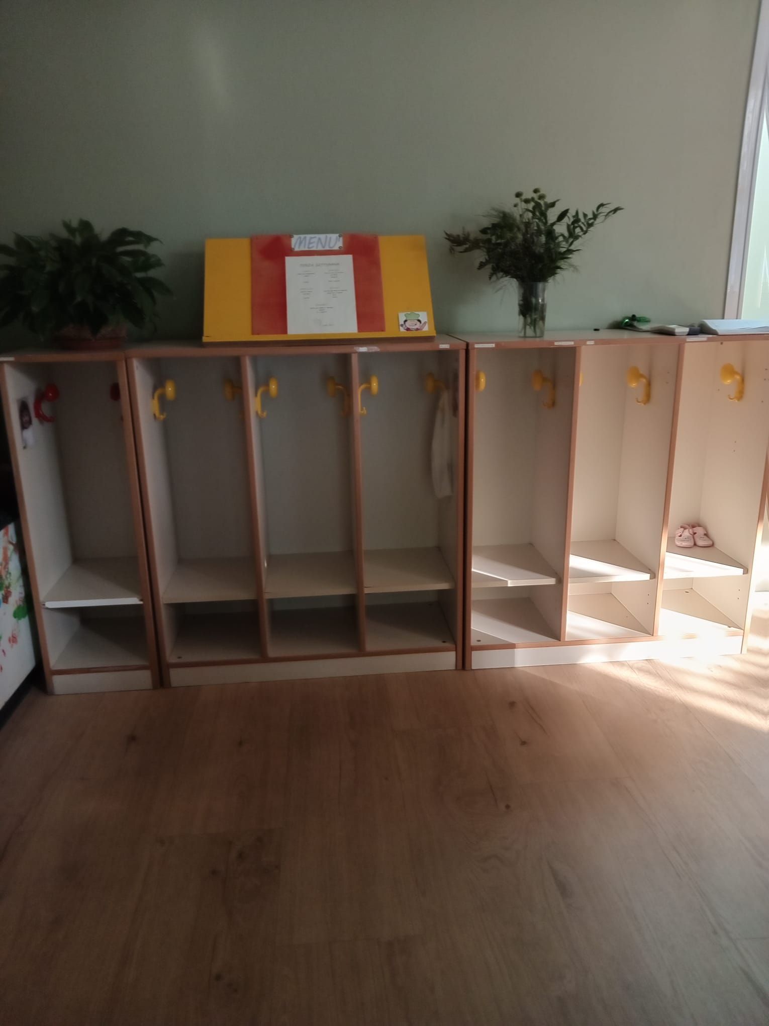 Armadietti in legno con ganci e ripiani contro una parete verde, su un pavimento in legno.