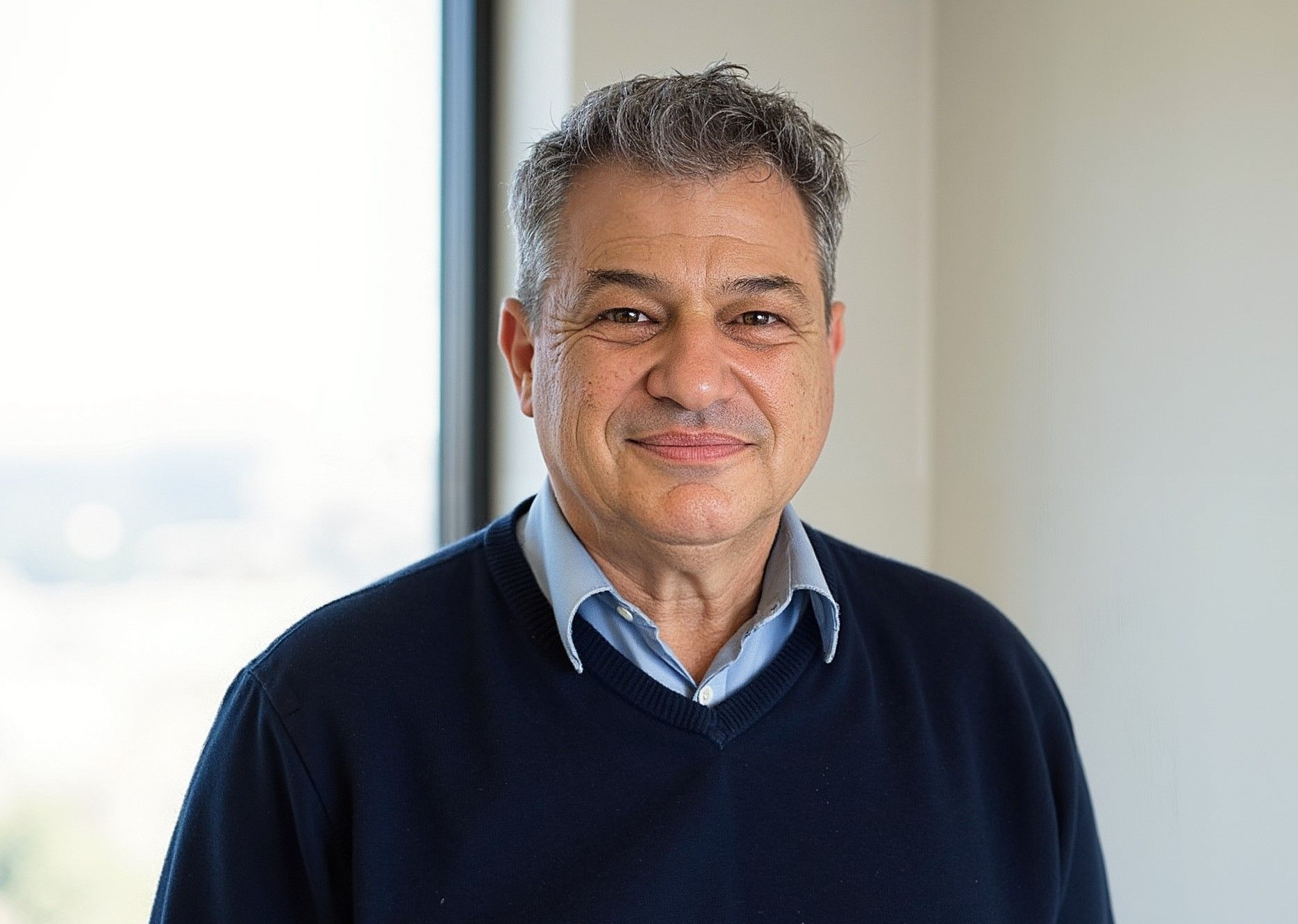 Man in a blue sweater and collared shirt smiles near a window.