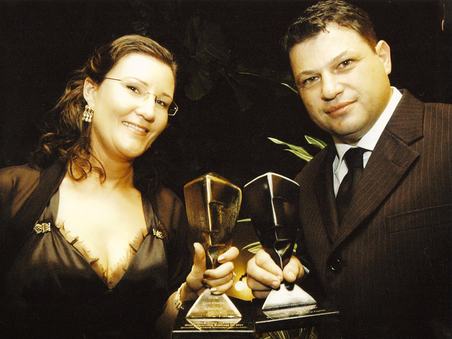 Woman and man holding trophies, smiling, formal attire.