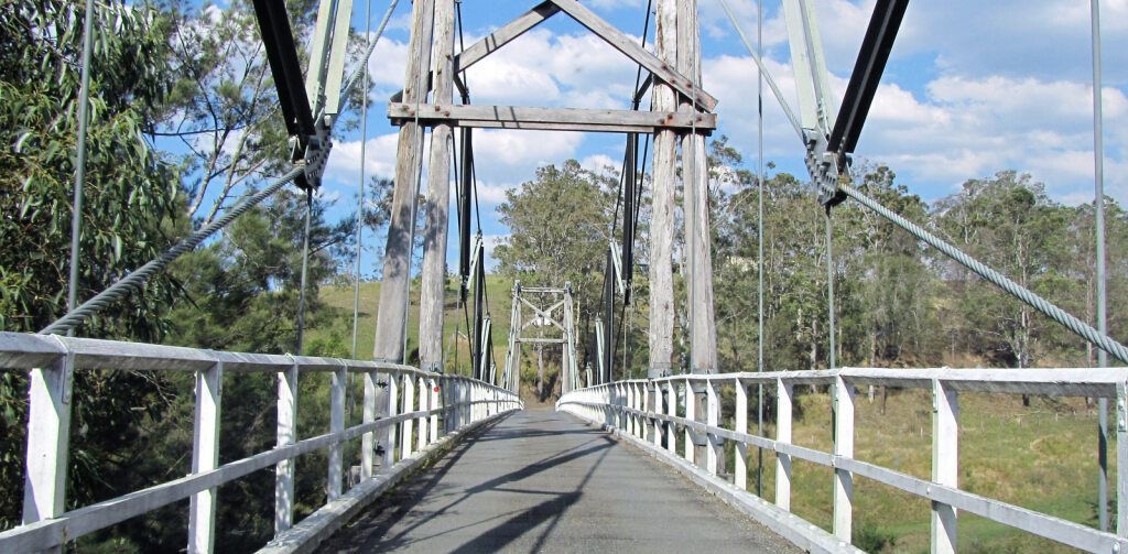 Suspension Bridge, White Railing, Wooden Posts, Grey Asphalt, Trees, Blue Sky — NRS Scaffolding Pty Ltd in Wauchope, NSW