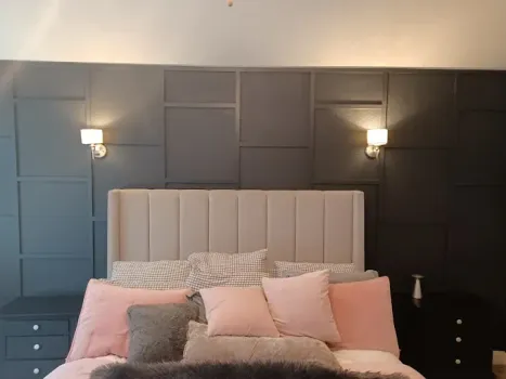 A bedroom features a charcoal panelled accent wall behind a beige headboard, soft pink pillows, and modern wall sconces.